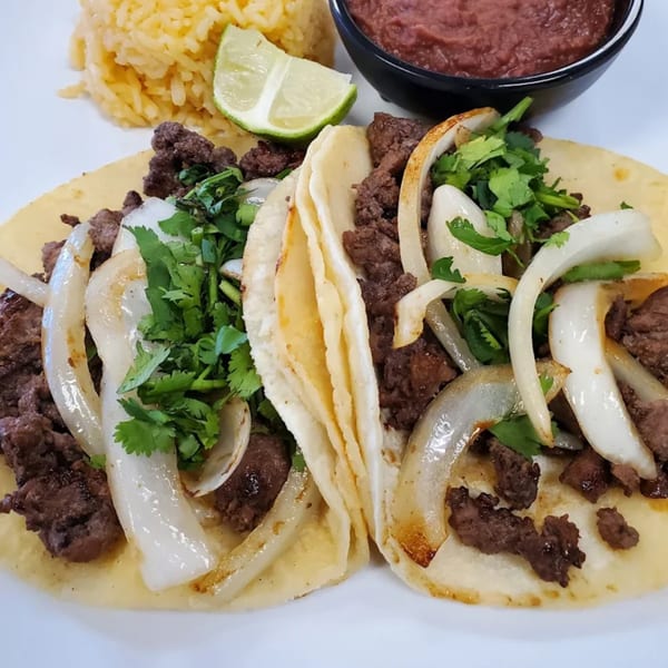 Two carne asada tacos with onions and cilantro, served with rice, beans, and a lime wedge.