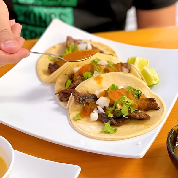 A hand spoons red salsa onto three El Sol Restaurant tacos with meat, onions, and cilantro.